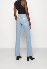 Helle blaue, ausgestellt geschnittene Jeans mit hoher Taille, die eine sichtbare Gesäßtasche und ausgeprägte Nähte aufweisen. Kombiniert mit schwarzen Stiefeletten.