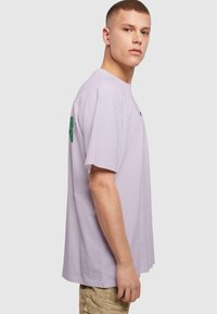 Camiseta de algodón de color púrpura claro con mangas cortas, que presenta un texto gráfico en verde en el brazo izquierdo y un corte relajado.