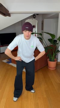 T-shirt branca com logótipo verde, calças pretas de pernas largas e sapatilhas prateadas. Gorro de malha bordô. Ambiente interior com plantas e mobiliário.