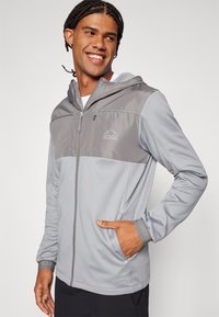 Siva dukserica s kapuljačom s patentnim zatvaračem na prednjoj strani, s tamnije sivim gornjim panelom i logotipom, izrađena od lagane tkanine s bočnim džepovima.