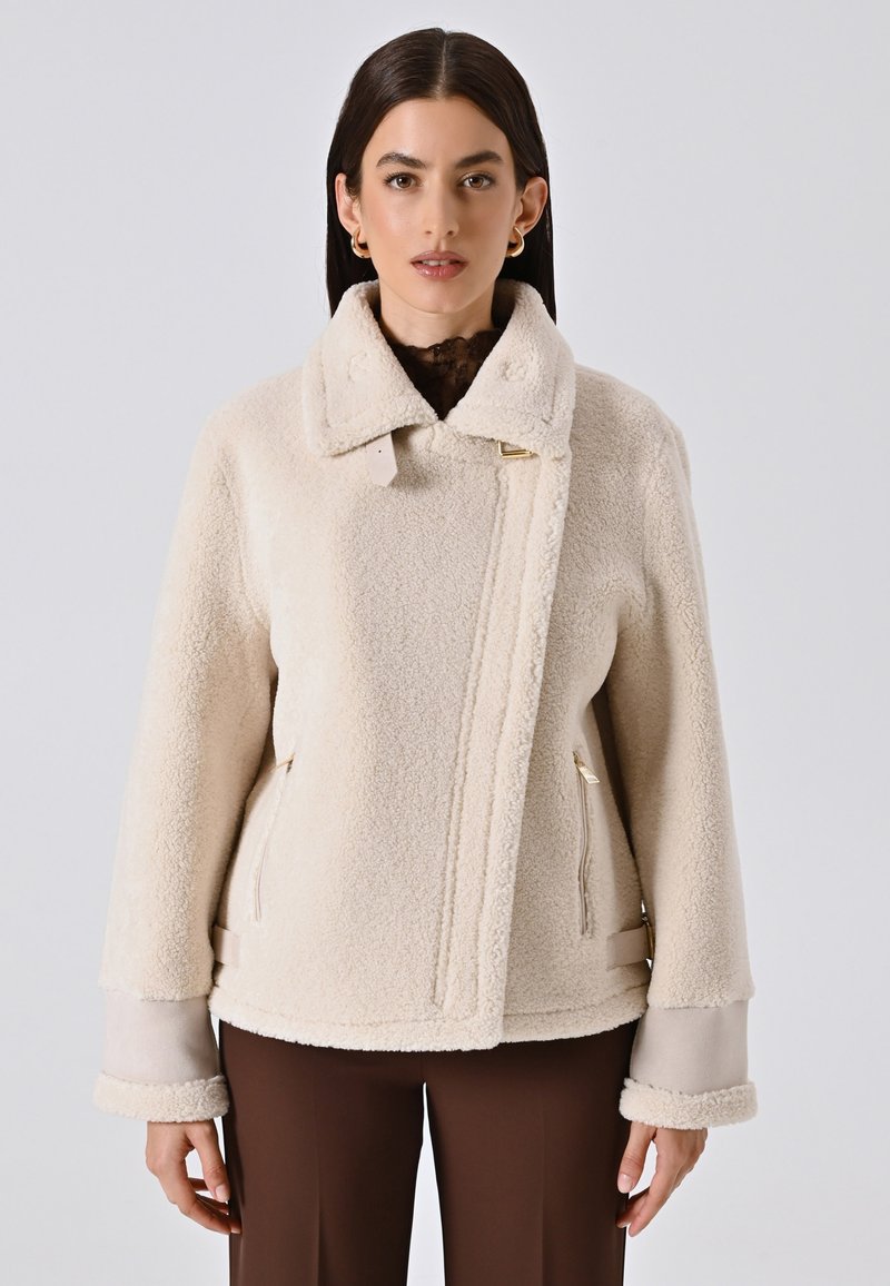 Giacca in pile color crema con colletto alto, tasche laterali e dettagli in metallo dorato. Presenta ampi polsini con una texture morbida.