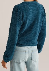 Femme portant un pull à manches longues texturé bleu canard foncé et un jean taille haute bleu clair, vue de dos sur un fond neutre.