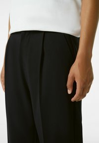 Pantalones plisados negros hechos de tela suave. La cintura está oculta debajo de una blusa blanca, con una mano apoyada junto al muslo.