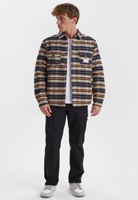 Camicia a quadri blu, marrone e crema con bottoni frontali, due tasche sul petto e un'etichetta. Abbinata a pantaloni neri e sneakers bianche.