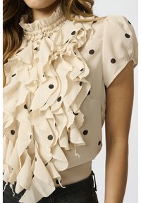 Beige blouse met zwarte stippen, met een gerimpelde voorkant en hoge hals. Korte mouwen en een getailleerd ontwerp. Zachte, gladde stof.