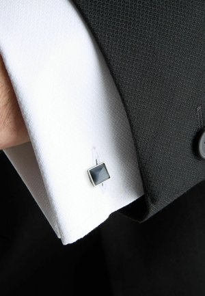 White textured dress shirt cuff with gray square cufflink partially covered by a black suit sleeve with a black button.