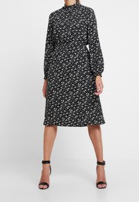 Svart klänning med långa ärmar, hög halsringning, åtsittande midja och vitt blommönster. Slät tyg, knälång, kombinerad med svarta klacksandaler.