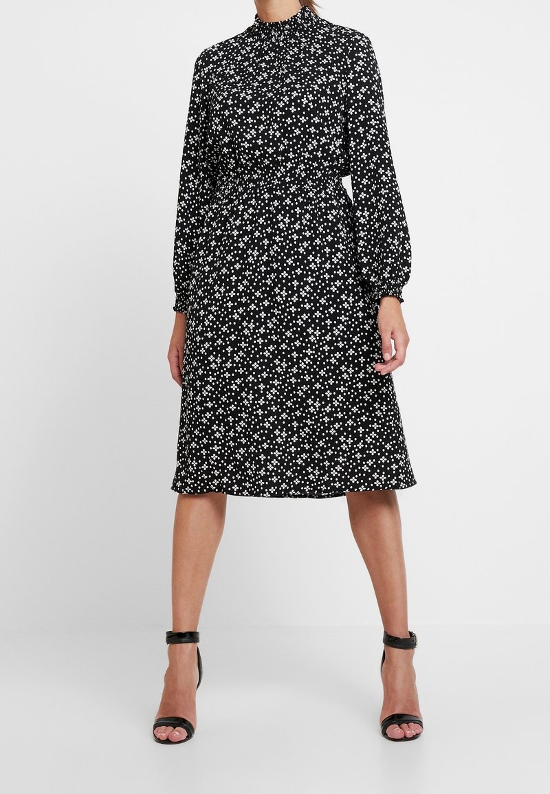 Svart klänning med långa ärmar, hög halsringning, åtsittande midja och vitt blommönster. Slät tyg, knälång, kombinerad med svarta klacksandaler.