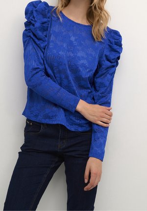 Vrouw draagt een blauwe bloemen kanten lange mouw top met opgepofte schouders en donkerblauwe spijkerbroek tegen een effen witte achtergrond.