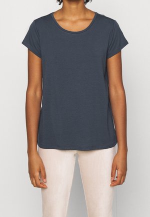 Frau trägt ein dunkelblaues T-Shirt mit kurzen Ärmeln und Rundhalsausschnitt sowie hellbeige, gerippte Hosen, steht vor einem einfarbigen Hintergrund.