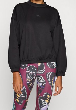 Persoon draagt een zwart topje met lange mouwen en een elastische zoom, en een legging met een meerkleurig vlinderpatroon tegen een effen achtergrond.