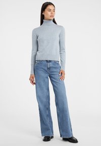 Maglione a collo alto blu chiaro in tessuto lavorato a maglia, abbinato a jeans a gamba larga in lavaggio chiaro. Stivaletti neri alla caviglia completano l'outfit.