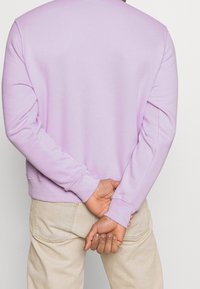 Sudadera lila de mangas largas, hecha de tela suave, con un cuello redondo y puños de canalé. Llevada con pantalones beige claro.