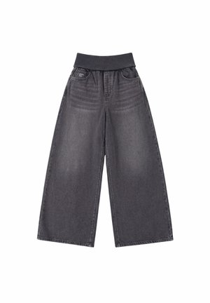 Modèle portant un jean noir délavé, ample et à jambes larges, avec une taille côtelée, photographié de la taille aux pieds sur un fond uni.