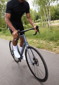 Vélo de route avec un cadre gris, des guidons noirs et des freins à disque. Le cycliste porte un maillot noir et des chaussures de cyclisme blanches sur un chemin pavé.