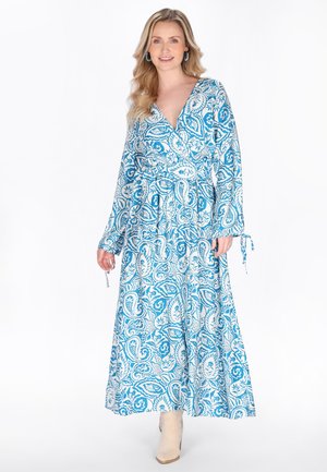 Donna bionda che indossa un lungo vestito avvolgente a motivi floreali blu e bianchi con maniche lunghe e stivaletti beige, sorridente.