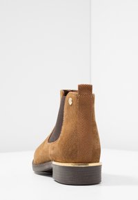 Stivale Chelsea in suede marrone con pannello elastico laterale, linguetta sul tallone, dettaglio dorato sopra la suola scura impilata, su sfondo bianco.