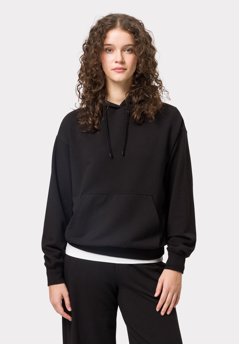 Černá mikina s kapucí vyrobená z měkké tkaniny, s přední kapsou, stahovací kapucí a žebrovanými manžetami. Nositá přes odpovídající černé kalhoty.