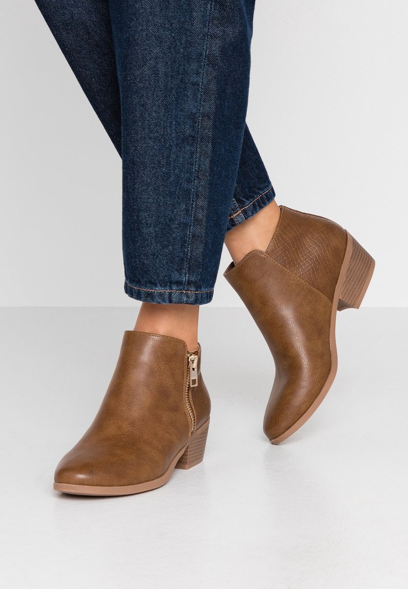 Bottes chevilles en similicuir marron avec fermeture éclair latérale, panneau arrière texturé et petit talon empilé, présentées avec un jean foncé.