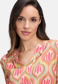 Femme aux longs cheveux noirs portant un haut coloré à motifs roses, jaunes et verts, regardant vers l'avant avec une expression neutre.