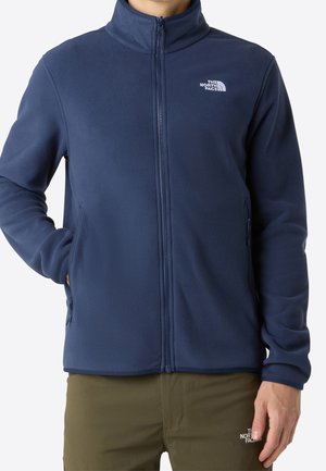 Mężczyzna w granatowej polarowej kurtce zapinanej na zamek i oliwkowych spodniach, oba z logo The North Face, stoi z jedną ręką w kieszeni.