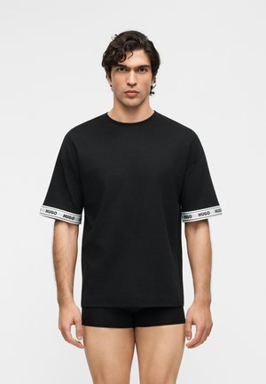 Hombre con cabello rizado y oscuro, vistiendo una camiseta negra extragrande con bandas blancas con la marca "HUGO" en las mangas y calzoncillos tipo bóxer negros.