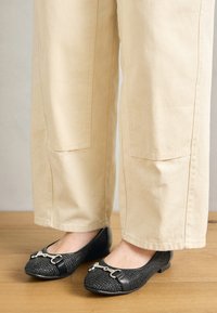 Scarpe nere a pianta piatta con una parte superiore texturizzata, rifiniture in pelle e accenti in metallo argentato. Abbinato a pantaloni beige a gamba larga.