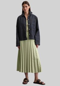 Denim jacket in dark blue with button closure and two front pockets, paired with a pleated, ankle-length light green dress and brown sandals.