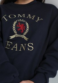 Marinblå sweatshirt med guld broderad text "TOMMY JEANS" och en sköldlogotyp med en röd drake inom en lagerdesign.