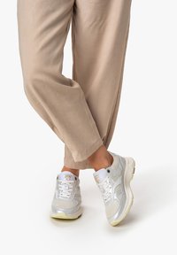 Valkoiset urheilukengät, joissa on hopeisia yksityiskohtia, verkkomateriaalia ja synteettistä päällistä, paksu pohja ja nauhakiinnitys, yhdistettynä beigeihin rentoihin housuihin.