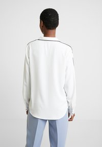Persona con capelli ricci corti che indossa una camicia bianca a maniche lunghe con bordature nere sulle spalle e pantaloni azzurri chiari, in piedi con la schiena rivolta.