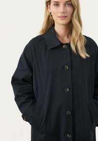 Námořnický modrý trench coat s límečkem, velkým knoflíkovým zapínáním, bočními kapsami a raglánovými rukávy. Hladká, lehká textilie.