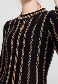 Maglione nero lavorato a maglia con motivo a catena verticale color sabbia, dotato di scollatura a girocollo e maniche lunghe, abbinato a una collana con pendente dorato.