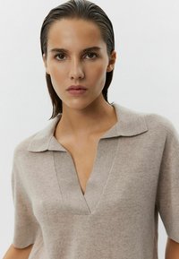 Béžový úpletový polo tričko s výstrihom do V a krátkymi rukávmi. Mäkká textúra a voľný strih. Minimalistický dizajn bez viditeľných vzorov alebo akcentov.