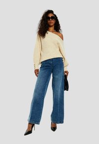 Maglione beige a maglia con design scollato, abbinato a jeans di denim blu a gamba larga con dettagli in bottone. Inclusi borsa nera e tacchi.