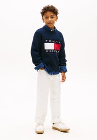 Μπλε πλεκτό πουλόβερ με κεντημένο λογότυπο, λεπτομέρειες σε λευκό και κόκκινο, συνδυασμένο με λευκό παντελόνι με γυρισμένα μπατζάκια και λευκά αθλητικά παπούτσια με ταμπά σόλες.