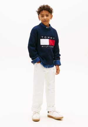 Pulover tricotat bleumarin cu o patch-logo, accente albe și roșii, asortat cu pantaloni albi suflecați și adidași albi cu tălpi bej.