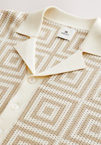 Beige gemustertes Polohemd mit geometrischen Designs, leichtem Strickstoff, zwei weißen Knöpfen und klassischem Kragen. Markenlabel sichtbar.