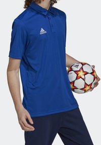 Polo de sport bleu avec col, en tissu lisse, arborant un logo Adidas blanc. Tenant un ballon de football décoré d'étoiles colorées.