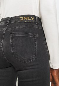 Jeans de denim negro con un diseño ajustado, que cuentan con un bolsillo trasero y una etiqueta de marca visible con letras doradas en la cinturilla.