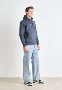 Donkerblauwe hoodie met een kangoeroezak, een capuchon met trekkoord en minimale tekst. Gepaarde met lichtblauwe wijde jeans en zwart-witte sneakers.