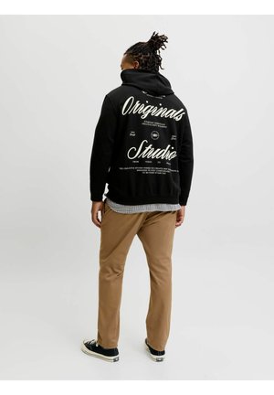 Person med dreadlocks iført sort hættetrøje med hvid "Originals Studio" tekst, khakibukser og sorte sneakers, stående mod en ensfarvet baggrund.