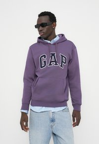Sweat à capuche violet avec logo "GAP" en blanc, poche kangourou à l'avant, associé à une chemise rayée bleu et blanc et un jean en denim clair.