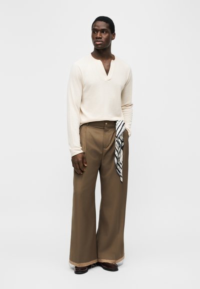 Uomo che indossa una camicia crema a maniche lunghe, pantaloni larghi marrone con un tessuto a righe appeso dalla tasca e scarpe marrone scuro, in piedi contro uno sfondo bianco.