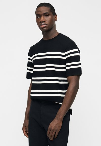 Άνδρας που φοράει μαύρο πουλόβερ με κοντά μανίκια και οριζόντιες λευκές ρίγες, σε συνδυασμό με μαύρο παντελόνι, στέκεται μπροστά σε φωτεινό φόντο.