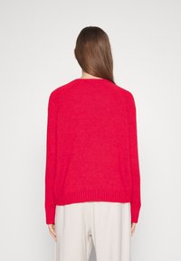 Roter gestrickter Pullover mit Rundhalsausschnitt, lockerer Passform, gerippten Säumen und strukturiertem Stoff. Getragen über hellen Hosen.