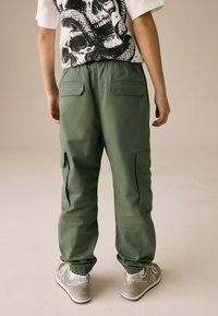 Pantalons cargo verts avec des poignets élastiques, dotés de plusieurs poches et d'une texture lisse. Portés avec des baskets grises.
