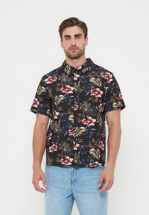 Άνδρας που φοράει μαύρο πουκάμισο με κοντά μανίκια, κόκκινο και μπεζ λουλουδάτο σχέδιο και ανοιχτό μπλε τζιν, στέκεται μπροστά σε λευκό φόντο.