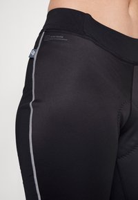 Czarne legginsy sportowe o gładkiej fakturze, z szarymi szwami bocznymi i regulowanym paskiem w talii. Zawierają metkę z logo w okolicy biodra.