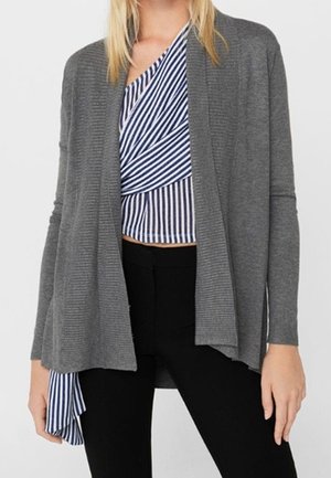Frau trägt eine graue offene Strickjacke über einem blau-weiß gestreiften Wickeltop und schwarzen Hosen, steht vor einem schlichten Hintergrund.
